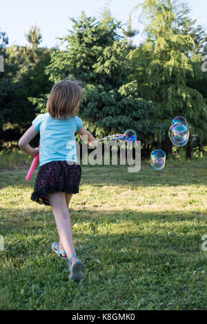 Giovane ragazza che gioca con la bacchetta di bolla in giardino, vista posteriore Foto Stock