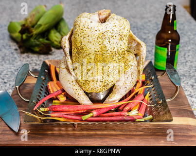 Un pollo intero montato su una lattina di birra padella per grigliare. pelato intero carote nel fondo della padella. bottiglia di birra e il coltello anche mostrato. Foto Stock