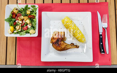 Pollo arrosto e sulla pannocchia di mais con una fresca insalata di verdure su un rosso tabella MAT. Foto Stock