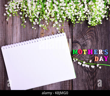 Vuoto bianco aperto il notebook e un elogiativo iscrizione con un giorno della madre su di un legno grigio Sfondo, nella parte superiore di un profumo di gigli di fioritura Foto Stock