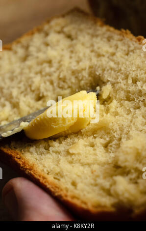 Chiusura del vero burro di essere spalmato su una fetta di pane appena sfornato, detenute dalla mano di sesso maschile con il pollice in frame. focaccia è appena visibile in background. Foto Stock