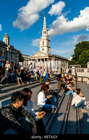 I giovani seduti sui gradini di Trafalgar Square con la chiesa di St Martin-in-the-Fields in backround, london, Regno Unito Foto Stock