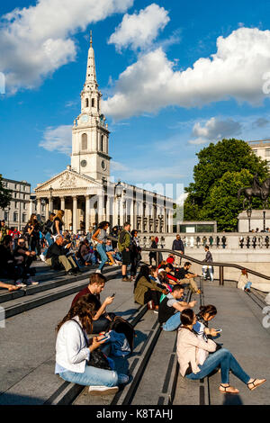 I giovani seduti sui gradini di Trafalgar Square con la chiesa di St Martin-in-the-Fields in backround, london, Regno Unito Foto Stock