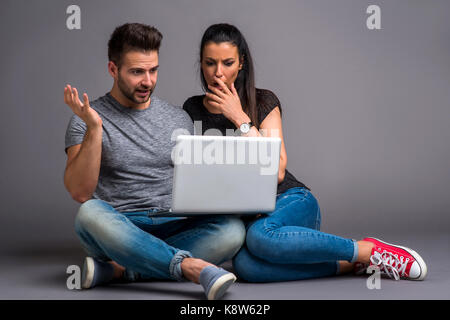 Una bella coppia giovane seduto per terra e guardando a un computer portatile e la sensazione di essere sorpreso Foto Stock