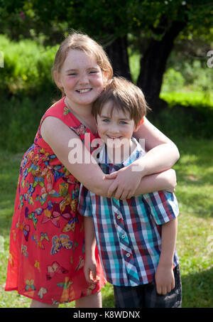 Un freckled dai capelli rossi ragazza abbraccia il suo pelo marrone fratello, sorridente al di fuori nel sole Foto Stock