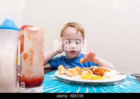 Un giovane ragazzo caucasico seduti a un tavolo a mangiare la sua cena con il ketchup bottiglia in primo piano Foto Stock