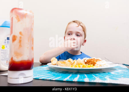 Un giovane ragazzo caucasico seduti a un tavolo a mangiare la sua cena con il ketchup bottiglia in primo piano Foto Stock