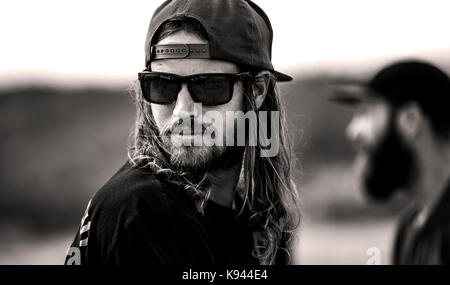Ritratto di uomo barbuto con i capelli lunghi, indossando occhiali da sole e cappello da baseball rivolto all'indietro. Foto Stock