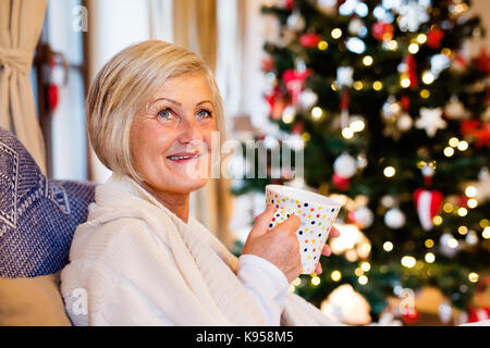 Senior bella donna seduta sul lettino di fronte illuminata albero di Natale all'interno nella sua casa godendo di tazza di tè o caffè, sorridente. Foto Stock