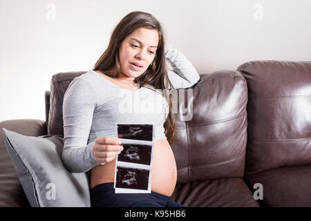 Donna incinta guardando il suo bambino di scansione ad ultrasuoni nel suo soggiorno Foto Stock