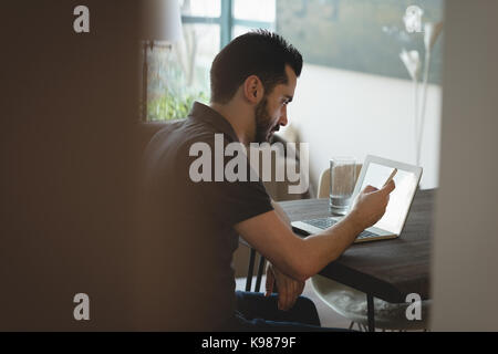 Uomo che utilizza il telefono cellulare mentre si lavora sul portatile a casa Foto Stock