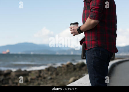 La sezione centrale dell'uomo con la tazza di caffè in piedi vicino alla spiaggia Foto Stock