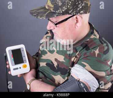 Senior anziano uomo indossa Abbigliamento camouflage controllare la sua pressione del sangue. monitor della pressione del sangue test nel suo braccio superiore. Foto Stock