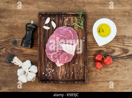 Crudo fresco di carni bovine taglio trasversale per l'ossobuco con spicchi di aglio, pomodori ciliegini, rosmarino, pepe, olio e sale sulla scheda servizio oltre rustico sfondo di legno Foto Stock