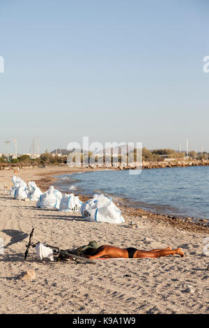 Un uomo è prendere il sole nella spiaggia di Glyfada (Atene, Grecia) che è inquinata da una fuoriuscita di petrolio. I sacchetti bianchi accanto all'uomo sono pieni di olio grezzo. Foto Stock