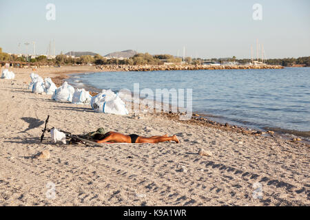 Un uomo è prendere il sole nella spiaggia di Glyfada (Atene, Grecia) che è inquinata da una fuoriuscita di petrolio. I sacchetti bianchi accanto all'uomo sono pieni di olio grezzo. Foto Stock