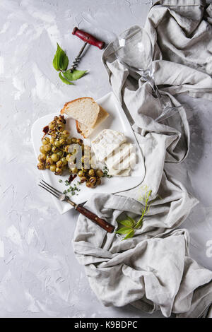 Piastra bianca con fette di formaggio camembert e cotto grappolo di uva verde servita con pane, bicchiere di vino, cavatappi, foglie verdi, forcella sul tessile su Foto Stock
