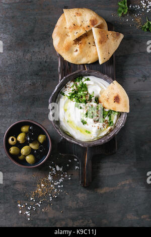 Labneh Middle Eastern libanesi di crema di formaggio dip con olio d'oliva, sale e le erbe aromatiche servita con olive, tradizionale pane pita nel recipiente di terracotta su te scuro Foto Stock