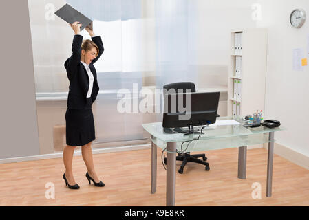 Arrabbiato imprenditrice permanente al desk gettando portatile in ufficio Foto Stock