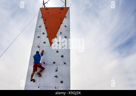 Persona che esercita la professione di estrema sportelderly scalatore maschio rende difficile spostare guardando in alto sul outdoor parete di arrampicata competizioni sportive. all'aperto sul cielo blu Foto Stock