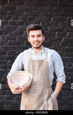 Potter, utensili, arte ceramica concetto - sorridente ceramista maschio con mani tenendo alcune bocce, carino prodotti di argilla e felice artigiano nel grembiule sullo sfondo scuro di un muro di mattoni, verticale. Foto Stock