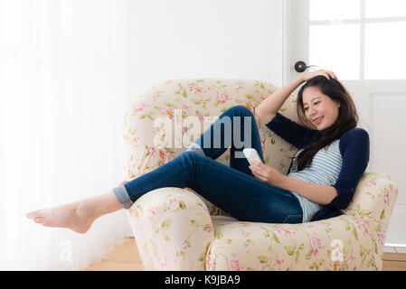 Elegante signora piacevole guardando mobile smartphone sensazione allegro e seduto sul divano di casa godendo di tempo di vacanza a casa. Foto Stock