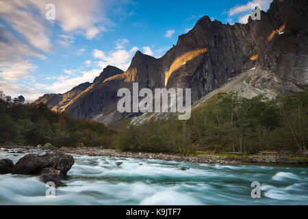 Al mattino presto nella valle di Romsdalen, Møre og Romsdal, Norvegia. In primo piano c'è il fiume Rauma e sullo sfondo c'è la parete verticale del Troll di 3000 piedi (in alto a destra nell'ombra), e le cime Trolltindane. La prima salita della ripida parete di Troll fu nell'estate del 1965. Una squadra inglese, e un'altra squadra norvegese hanno scalato il difficile muro attraverso due percorsi diversi. Foto Stock