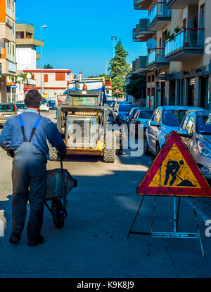 Lavori stradali di rosso e di giallo segno a triangolo sulla recinzione metallica. Strada in costruzione. lavoratore si muove carriola Foto Stock