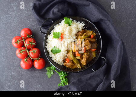 La carne di maiale piccante salsa al curry con melanzane e fagiolini, servito con riso cotto in un nero ciotola di metallo su un grigio Sfondo astratto. cucina casalinga. Foto Stock