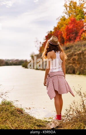 In autunno, armonia, concetto di viaggio. giovane ragazza vestita in abbigliamento sportivo in piedi sulla riva del fiume, sullo sfondo vi è la foresta di rosso e al giallo alberi come nel fuoco Foto Stock