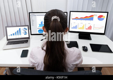 Vista posteriore di imprenditrice analizzando i grafici su più computer alla reception Foto Stock