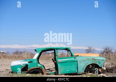 Abbandonato e arrugginito relitto di un vecchio verde sovietica vintage vettura russa nel mezzo di estratto secco di terreni agricoli con scenic montagne dalle cime ricoperte di neve e di clea Foto Stock