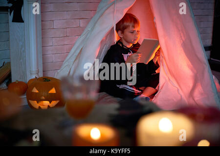 Concentrato ragazzo carino vestito come scheletro seduta all'interno di teepee e la lettura di libri interessanti da sola nella stanza buia durante la notte di Halloween Foto Stock