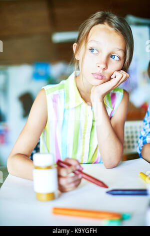 Ritratto di bella bambina fantasticando guardando lontano pensively durante la classe d'arte nella scuola, seduta con foglio di carta bianco Foto Stock