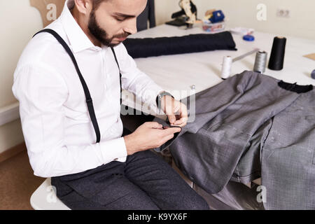 Talentuoso sarto barbuto seduto al tavolo di lavoro e la cucitura di custom-made giacca maschio da mani, interno del moderno studio sullo sfondo Foto Stock