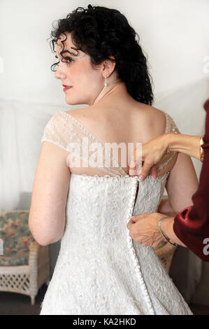 Bella Sposa si erge nel profilo mentre la madre zips abito nuziale. sposa ha a lungo, curling capelli neri e camice ha piccolo cappuccio maniche con bordatura e l Foto Stock