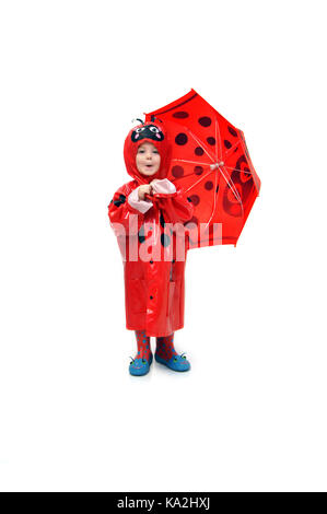 Adorabile bambina indossa un impermeabile e un ombrello rosso, ladybug. Sta facendo una faccia divertente e sta levandosi in piedi in una stanza tutta bianca. Foto Stock