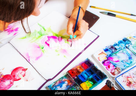 Un close-up artist pitture e vernici in un album per il disegno di una sottile spazzola in legno di rosa con colori ad acquerello, sul tavolo è una custodia in pelle con brus Foto Stock