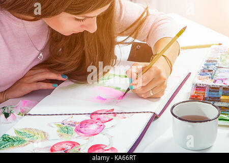 Dai capelli scuri donna giovane richiama una spazzola in legno e acquerello in un disegno di pad con peonie rosa con foglie verdi, sul tavolo si trovano una custodia in pelle con Foto Stock