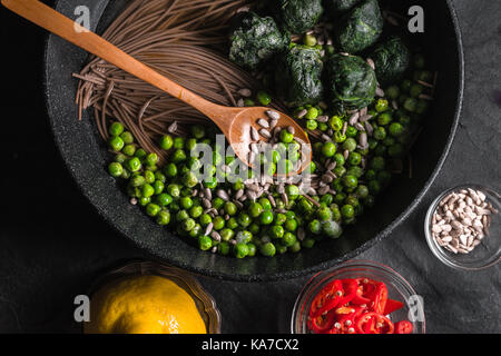 Tagliatelle di grano saraceno in una padella, piselli congelati e spinaci, peperoncini e semi in una ciotola in orizzontale Foto Stock