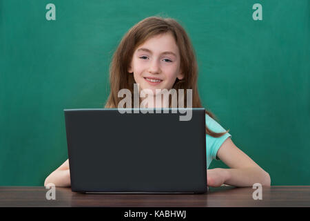 Felice ragazza utilizzando laptop al banco contro la lavagna in aula Foto Stock