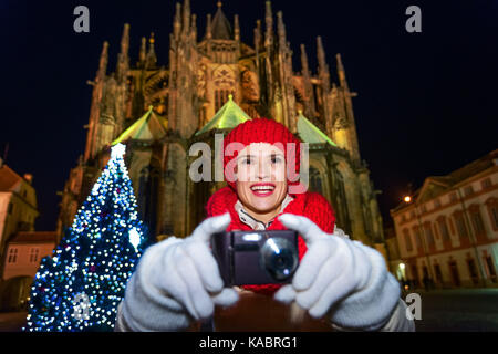 Magia sulle strade della città vecchia a Natale. Sorridendo viaggiatore moderno Woman in Red Hat e sciarpa nei pressi di albero di natale nella parte anteriore del st. vitus cathe Foto Stock