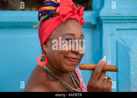 Un colorfully vestito donna cubana detiene un sigaro come si siede sul lato della strada nella habana vieja a l'Avana, Cuba. Foto Stock