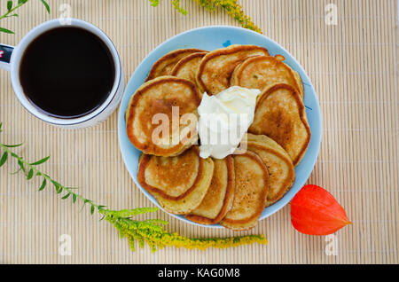Cucina casalinga - frittelle fritte con la farina di segale con panna acida e una tazza di caffè. decorato con fiori e physalis. sfondo di legno Foto Stock
