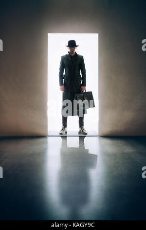 Silhouette uomo in hat e impermeabile in piedi la luce di apertura della porta in camera oscura Foto Stock