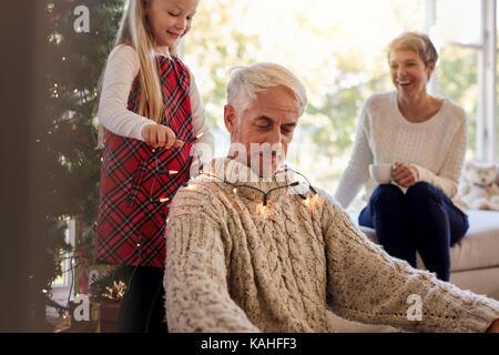 Bambina mettere le luci di Natale intorno a suo nonno il collo con la nonna seduta in background. Famiglia avente divertimento mentre la decorazione casa du Foto Stock