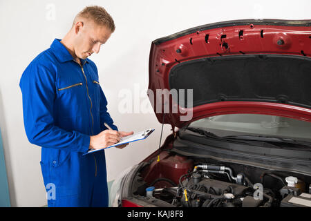 Mechanic in piedi accanto a auto sulla scrittura di appunti in garage Foto Stock