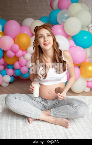 Giovane donna incinta bere latte seduta sul pavimento della camera. Foto Stock