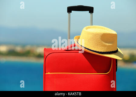 Estate hat sulla valigia rossa close-up. vacanza e concetto di viaggio. valigia e cappello giallo sul soleggiato mare sullo sfondo. Foto Stock