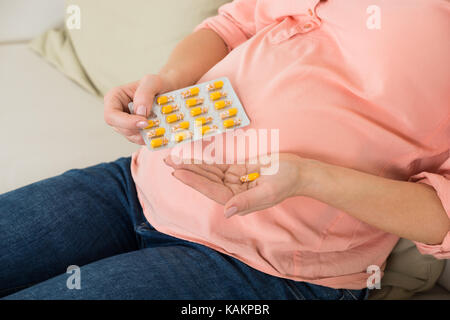 Sezione mediana della donna incinta prendendo vitamina pillola sul divano di casa Foto Stock